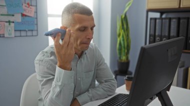 Genç adam ofisteki masasında çalışırken akıllı telefonundan sesli mesaj dinliyor, odaklanmış bir ifade ve modern çalışma alanı var..
