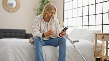 Uzun sarı saçlı İspanyol bir adam, yatak odasında oturuyor, akıllı telefon kullanıyor, ve yanında koltuk değnekleriyle bir kupa tutuyor..