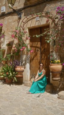 Yeşil elbiseli İspanyol kadın Valldemossa, Mallorca 'nın büyüleyici sokaklarında oturuyor. Etrafı güzel çiçekler ve geleneksel İspanyol taş mimarisi ile çevrili..
