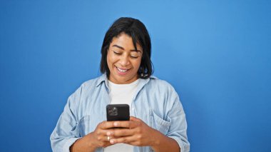 Gülümseyen bir İspanyol kadın telefonunu izole edilmiş mavi bir duvara bakıyor..