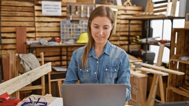 Gündelik giyinmiş genç bir kadın, iyi donanımlı bir marangozluk atölyesinde dizüstü bilgisayarına odaklanır..