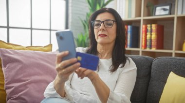 Olgun bir kadın rahat bir oturma odasında akıllı telefon ve kredi kartı kullanır. İnternetten alışveriş veya bankacılık gibi..