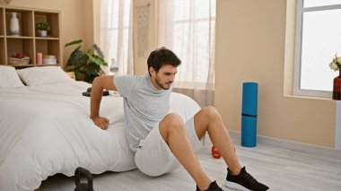 Güneşli, mobilyalı bir yatak odasında bir yatağın kenarında oturan, kettlebell ile egzersiz yapan, gündelik kıyafetli yakışıklı bir genç..