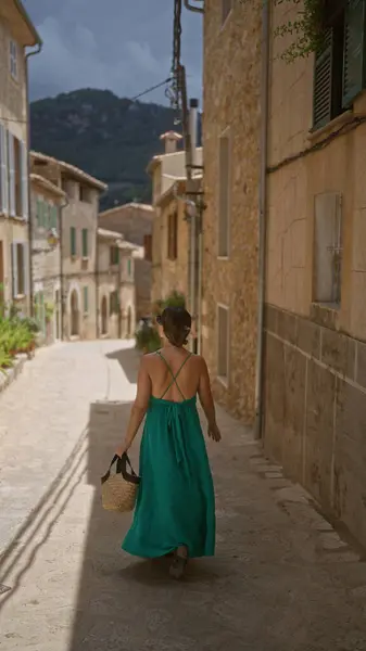 stock image Hispanic woman in a green dress walking through a charming narrow street in valldemossa, mallorca, spain, holding a wicker bag under a cloudy sky.