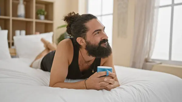 stock image A bearded young man relaxes in a bedroom with a coffee, embodying leisure and comfort in a casual setting.