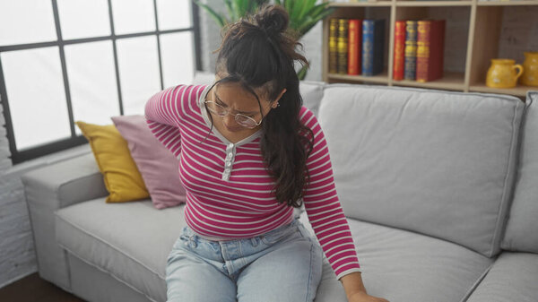 Латиноамериканка с болью в спине, сидящая на диване в помещении, выглядящая расстроенной