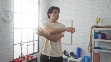 Yakışıklı genç bir adam kollarını kapalı bir spor salonunda geriyor. Ekipman dolu, gösterişli bir spor salonu ve kararlılık..