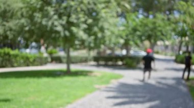 Güneşli bir günde yeşil ağaçlar ve yeşilliklerle çevrili bir parkta kıvrımlı bir yolda yürüyen adamların bulanık görüntüsü.