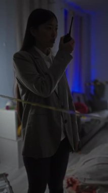 Asyalı bir kadın telsizle iletişim kurarken loş ışıkta yanan bir suç mahallini araştırıyor.