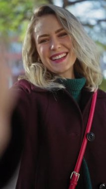 Sarı saçlı, genç, mutlu, beyaz bir kadın, bordo bir ceket giyiyor, yemyeşil bir şehir parkında canlı bir açık hava portresi için poz veriyor..