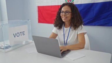 Seçim odasında dizüstü bilgisayarı ve oy kutusu olan olgun bir kadın Dominik Cumhuriyeti bayrağıyla süslenmiş.