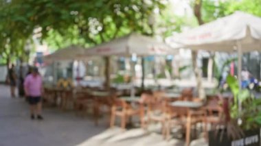 Güneşli bir günde şehir parkında masaları ve şemsiyeleri çözünmüş bir kafenin yanında yürüyen bir adam.