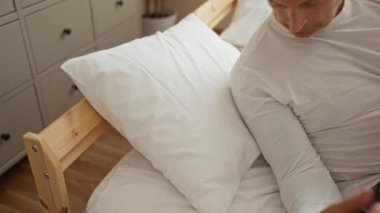 Beyaz şezlonglu orta yaşlı bir adam gündüzleri rahat bir yatak odasında uyuyor. Tahta yatak ve beyaz yastıklı.