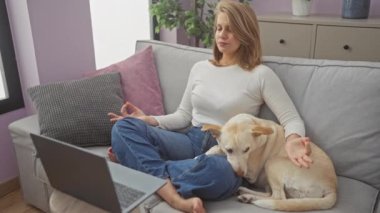 Genç bir kadın rahat bir dairede dizüstü bilgisayarı ve köpeği ile kanepede meditasyon yapıyor.