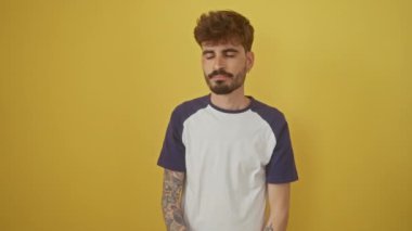 Genç İspanyol adam, izole edilmiş sarı bir arka planda duruyor, yorgun gözleri ve burnu ovalıyor, baş ağrısı, yorgunluk, stres, hüsran ve baş ağrısı belirtileri gösteriyor.