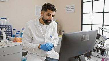 Laboratuvar önlüğü ve eldiveni giyen genç bir İspanyol parlak bir laboratuvardaki bilgisayarda dikkatlice çalışıyor..