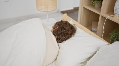 A curly-haired man sleeping in a bedroom with white bedding and a wooden headboard, portraying a relaxed, comfortable indoor setting. clipart