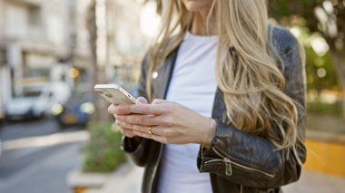 Sarışın genç bir kadın güneşli bir şehir sokağında akıllı telefon kullanıyor. Şehirde modern bağlılık ve günlük yaşam tarzını somutlaştırıyor..