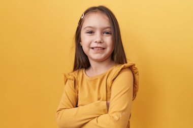 Tapılası İspanyol kız, özgüven ve mutluluğun resmi, sarı izole bir arka planın önünde duruyor. Kollarını kavuşturdu, parlak bir şekilde gülümsüyor, doğrudan kameraya bakıyor..