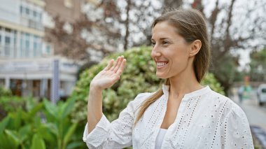 A smiling young woman in a white blouse greets someone on a sunny city street, portraying casual elegance and friendliness. clipart