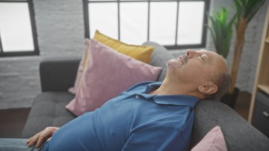 Orta yaşlı bir adam, modern bir oturma odasındaki kanepede huzur içinde uyuyor. Rahatlık ve rahatlık sergiliyor..