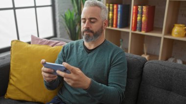 Modern oturma odasında renkli yastıklar ve kitaplık ile akıllı telefon kullanan orta yaşlı, beyaz sakallı bir adam..