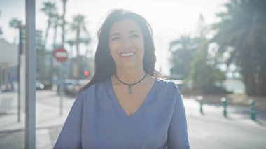 Gülümseyen orta yaşlı İspanyol kadın güneşli bir caddede dışarıda duruyor.