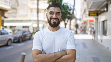 Genç sakallı bir adam güneşli bir caddede kollarını kavuşturup gülümsüyor..
