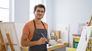 Parlak bir sanat stüdyosunda resim yapan yakışıklı, gözlüklü ve önlüklü, elinde renkli boyalı bir palet tutan bir İspanyol..
