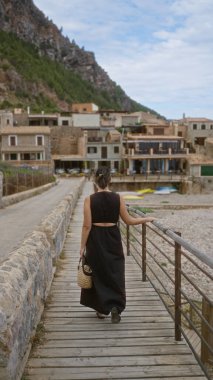 Port de valldemossa, mallorca, İspanya 'da tahta bir patikada yürüyen bir kadın. Bulutlu bir günde arka planda taş binalar ve dağlar var.