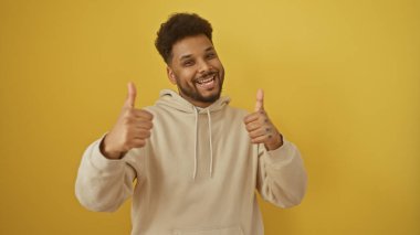 Gülümseyen Afro-Amerikan adam başparmağını kaldırıyor, sarı bir arka planda kapüşonlu giyiyor..