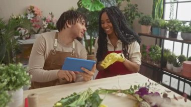 Çiçekçi bir adam ve kadın, arka planda bitkiler olan bir çiçekçide tablet kullanırken gülümsüyor..
