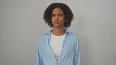Genç Afro-Amerikalı kadın gömleğini giyiyor. Kafası karışık, kafası karışık, soyutlanmış beyaz arka plan üzerinde şüpheli bir yüz ifadesi var. Belirsiz bir hareketle merak ediyor ve omuz silkiyor.
