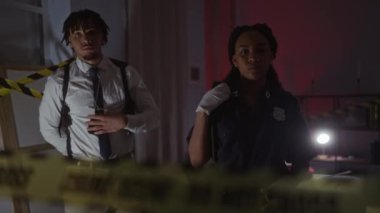 Polis gibi giyinmiş bir kadın ve bir erkek, sarı bant, el feneri ve ciddi ifadelerle gece suç mahallinde dururlar..
