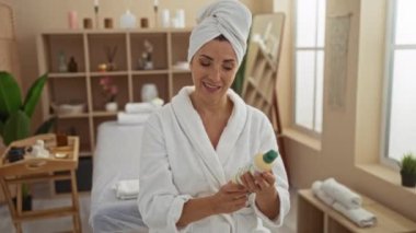 Ürünleri arka planda bitki ve raflarla dolu rahat bir spa ortamında tutan kadın, kafasında sabahlık ve havlu, kapalı alanda sakin ve şımartıcı bir seans olduğunu ima ediyor.