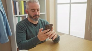 Sakallı, akıllı telefon kullanan, evde rahat bir yaşam tarzını canlandıran yakışıklı, olgun bir adam..