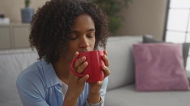 Kıvırcık saçlı genç Afrikalı Amerikalı kadın evdeki rahat oturma odasında sıcak bir içecekten zevk alıyor, rahatlığı ve rahatlığı yansıtıyor..