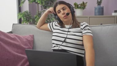 Çizgili tişörtlü, kulaklık takan genç bir İspanyol kadın evde, gri bir kanepede dizüstü bilgisayarla çalışırken yorgun görünüyor..