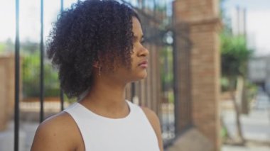 Kıvırcık saçlı, güzel bir Afro-Amerikan kadını, beyaz kolsuz bir bluz giyiyor, arka planında demir çit ve tuğla sütunlar olan şehir sokaklarında dikiliyor..