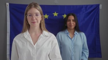 Avrupa bayrağı önünde gülümseyen iki kadın basit bir iç mekanda birlik ve çeşitliliği somutlaştırıyor.