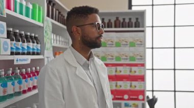 Kendine güvenen genç bir adam ilaç raflarıyla dolu modern bir eczanede eczacı olarak duruyor..