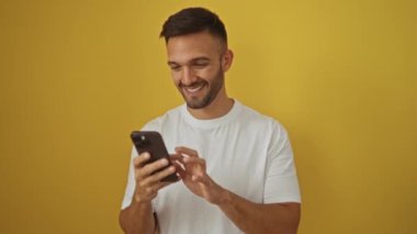 Genç İspanyol adam akıllı telefon kullanıyor, tamam işareti var, gülümsüyor ve işaret ediyor sarı arka planda, kendinden emin ve mutlu.