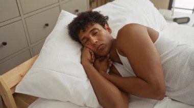 Yatak odasındaki bir yatakta yatan genç adam düşünceli ve düşünceli görünüyor. Etrafı yastık ve mobilyalarla çevrili ve beyaz bir atlet giyiyor..