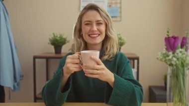 Yeşil kazaklı neşeli sarışın bir kadın, sıcak bir evin içinde kahvenin tadını çıkarıyor..