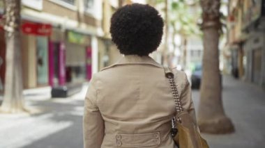 Orta yaşlı, kıvırcık saçlı, sırıtan bir kadın. Sokakta bej bir ceket giyerek yürürken kollarını kavuşturuyor.