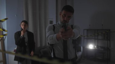 Profesyonel kıyafetli bir adam ve bir kadın loş ışıklı, kapalı bir suç mahallinde, birinin elinde silah, diğerinin ise telsiz kullanarak ayakta duruyorlar..