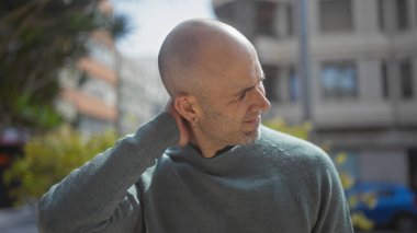 Sakallı ve sakallı genç bir adam güneşli bir şehir sokağında boynunu tutuyor, erkeklik saçıyor..