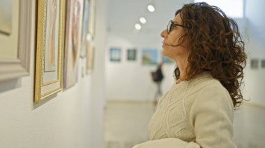 Gözlüklü olgun İspanyol kadın kapalı bir sanat galerisindeki resimlere hayran..