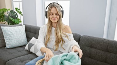 Genç bir kadın modern bir oturma odasında çamaşır katlarken kulaklıkla müzik dinlemekten hoşlanır..