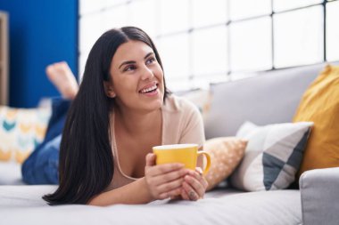 Young beautiful hispanic woman drinking coffee lying on sofa at home